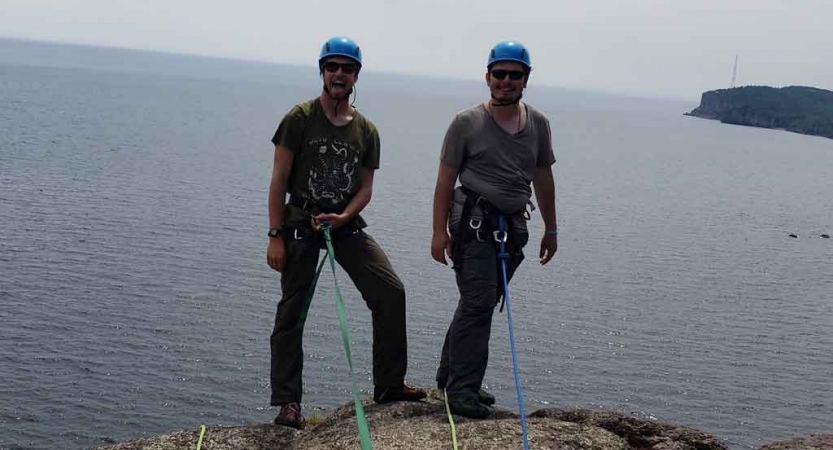 two students secured by ropes stand on a cliff overlooking a blue lake and prepare to rock climb